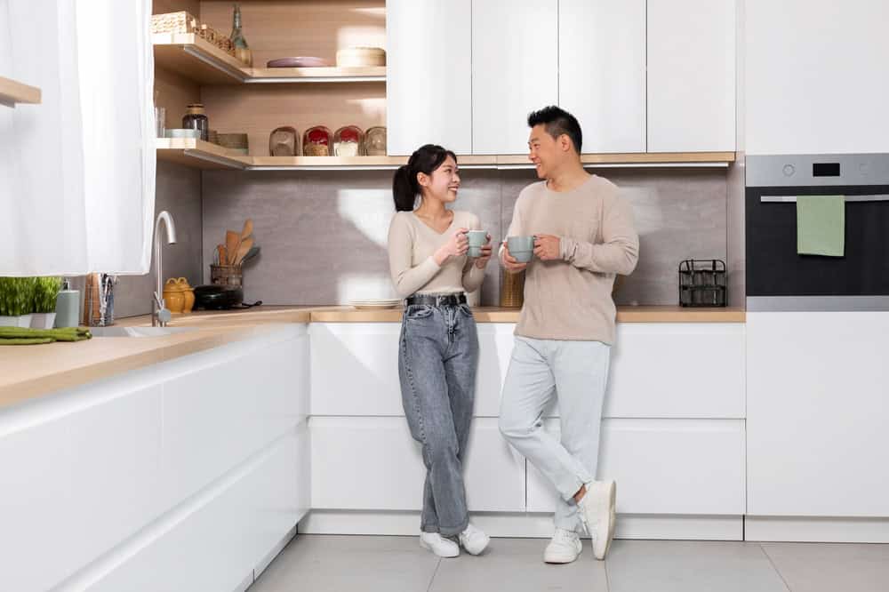 A couple smiles at each other while standing in a modern kitchen. Both hold mugs and are dressed in casual, neutral-colored clothing. The kitchen features white cabinets, wooden shelves, and various kitchenware.